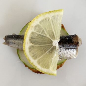 Image of a Yurrita Sardinillas (Small Sardines) in Extra Virgin Olive Oil 10/14 on a cracker with a round of cucumber and a thinly sliced lemon half