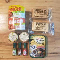 Image of RTG bento bag contents including Mini Tabasco hot sauces, mustards, Sempio garlicleaves, crackers, and Sempio kimchi